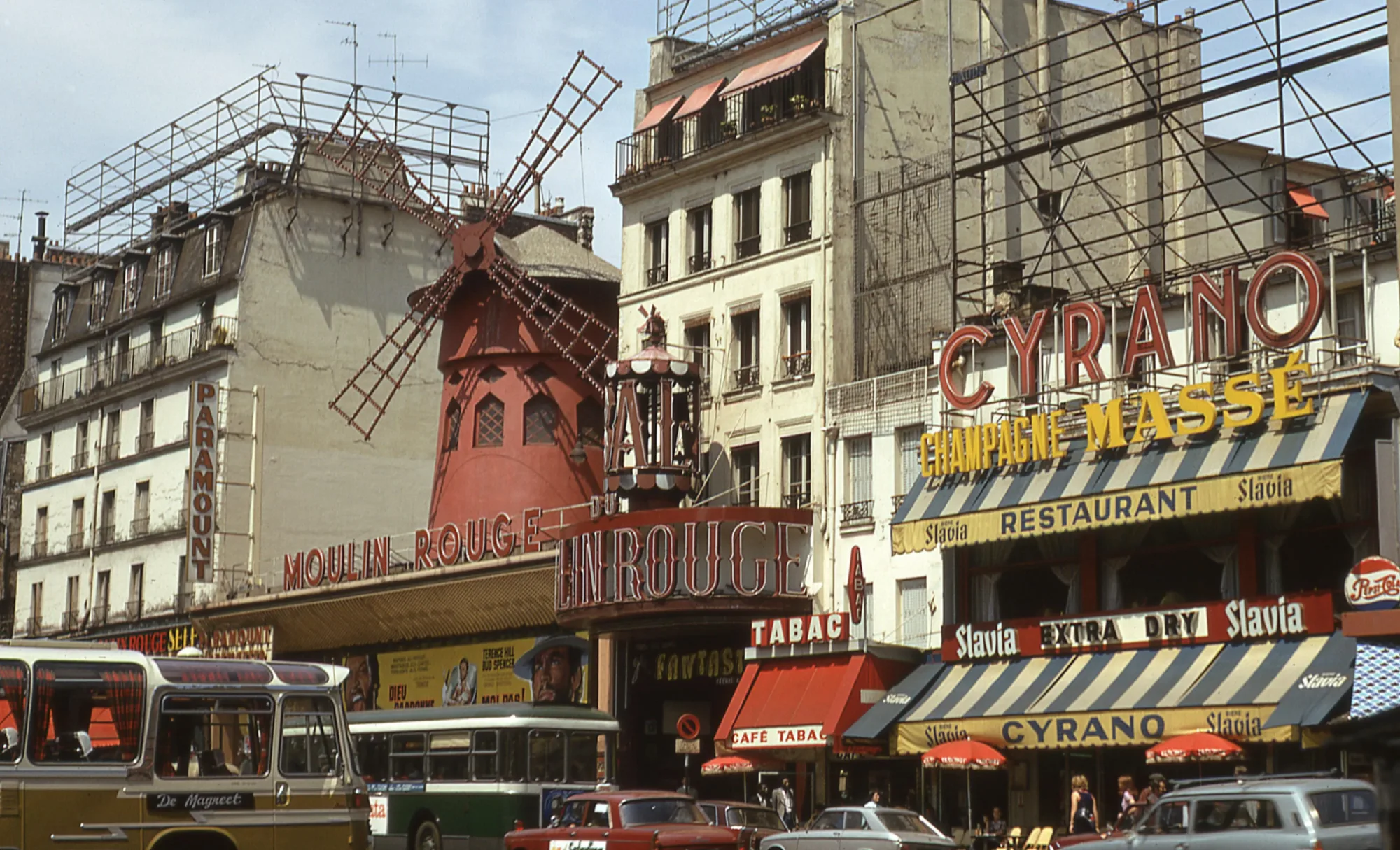 Photographie vintage d'une rue parisienne dans les années 1970 devant le Moulin Rouge, avec bus, voitures aux teintes vives et enseignes colorées Cyrano et Champagne Massé