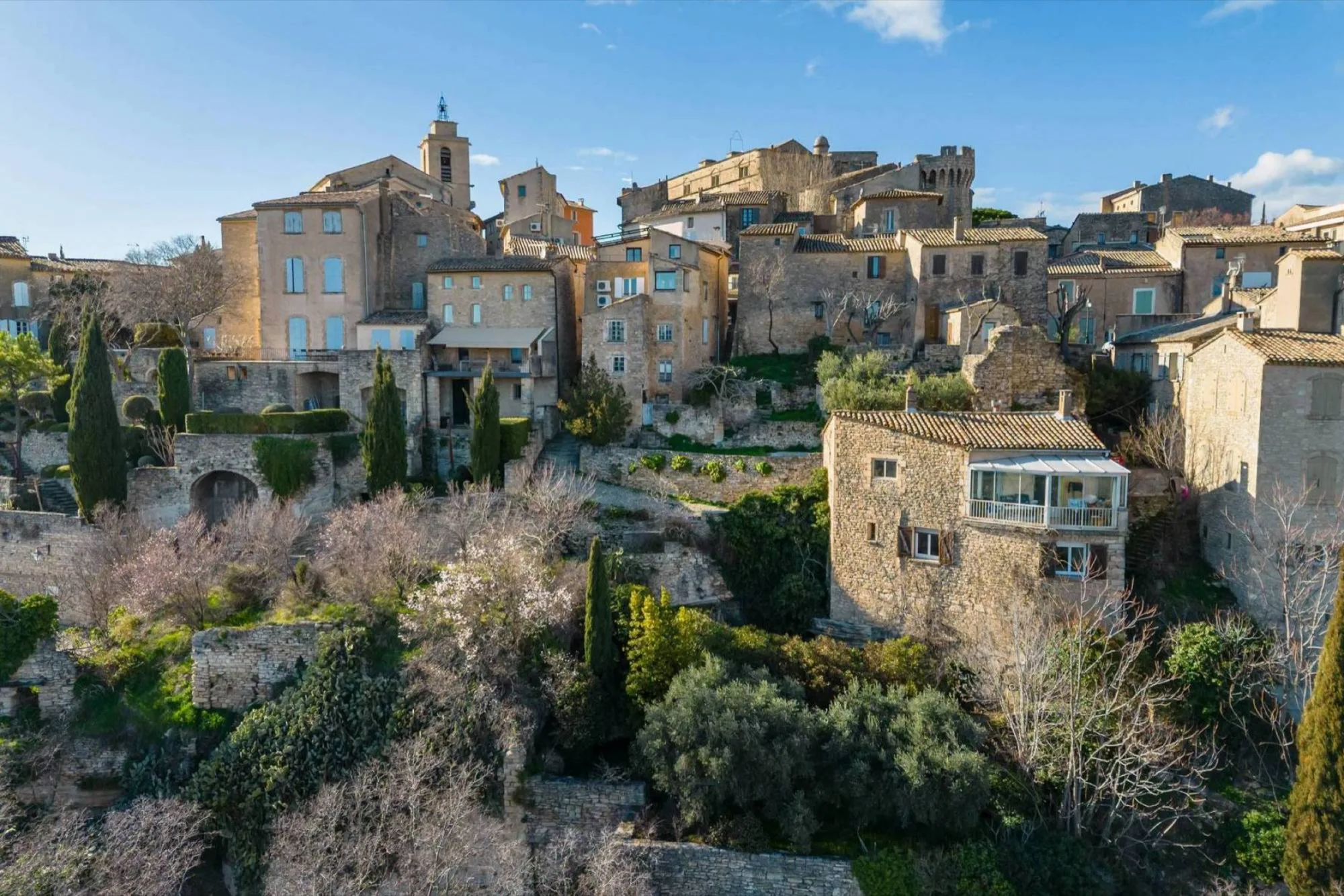 Vue aérienne d'un village perché du Luberon en pierre calcaire ocre — maisons médiévales serrées, cyprès et clocher se détachant sur un ciel bleu