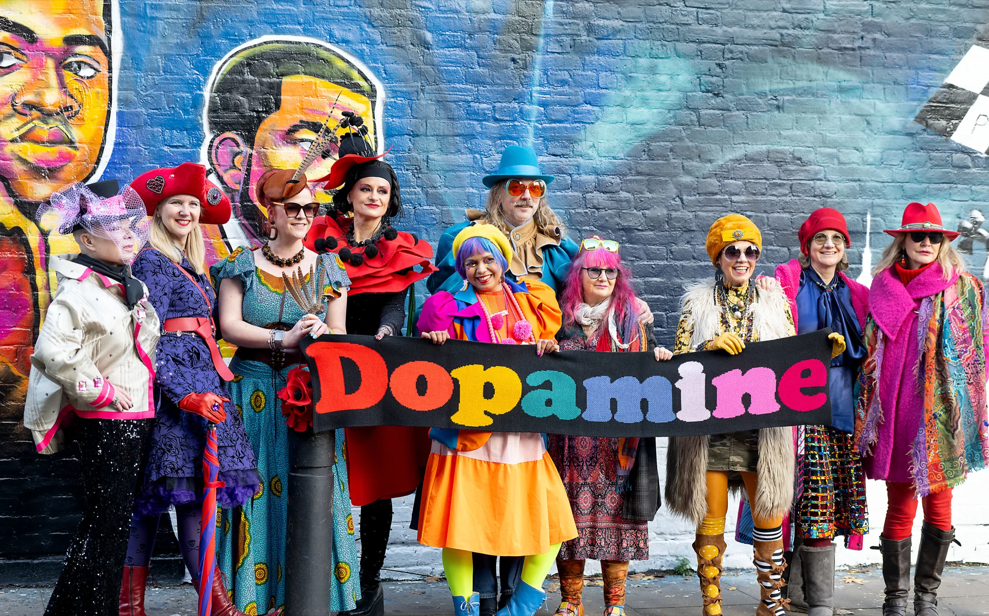 Groupe de femmes en tenues très colorées — robes à motifs, chapeaux vermillon et bleus, écharpes roses — tenant une banderole tricotée « Dopamine » devant une fresque murale urbaine