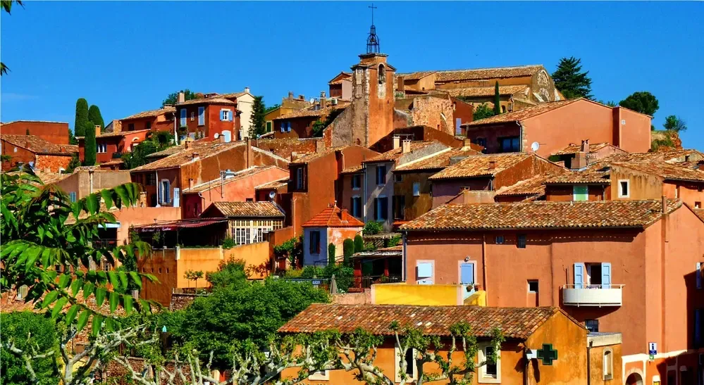 Village de Roussillon dans le Luberon — maisons en ocre rouge et orange étagées à flanc de colline sous un ciel bleu, illustration vivante d'un chromatisme régional préservé